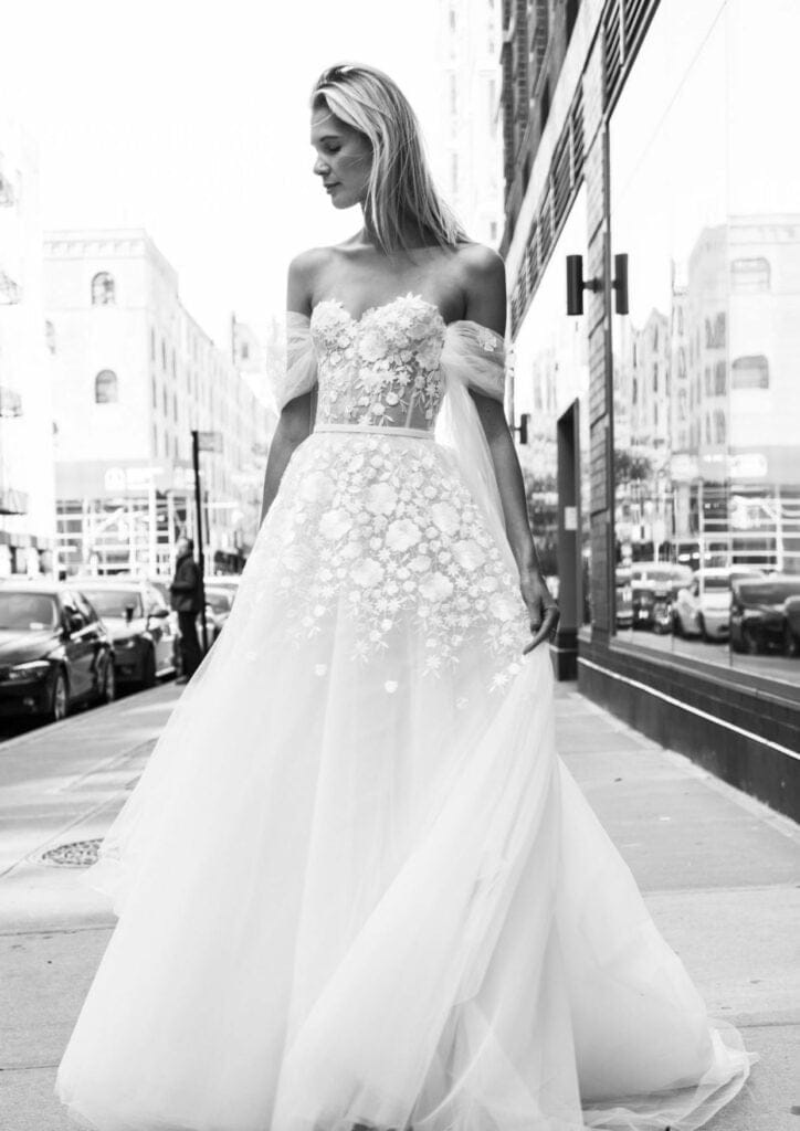 Bride in floral gown on city sidewalk, monochrome.