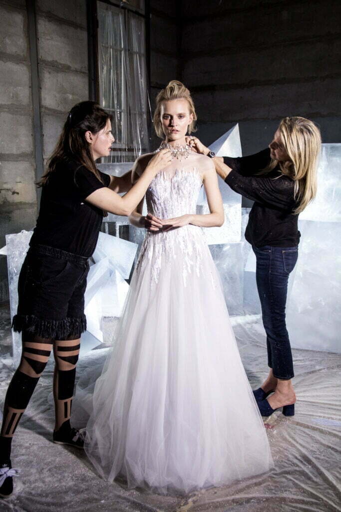 Model in white gown, stylists adjust hair.