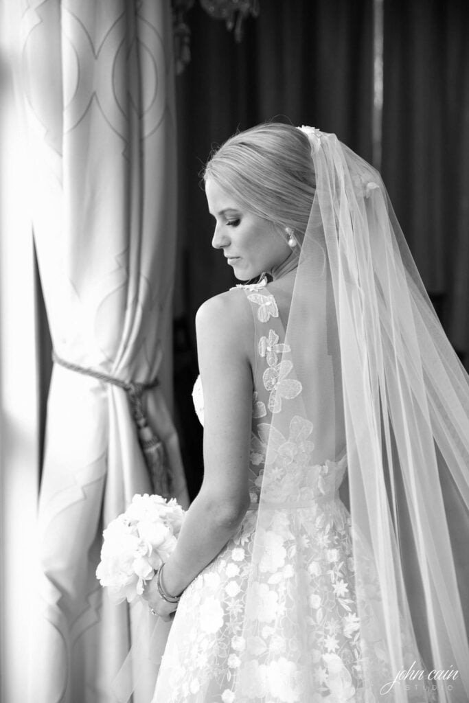 Bride by window, holding bouquet, softly lit.