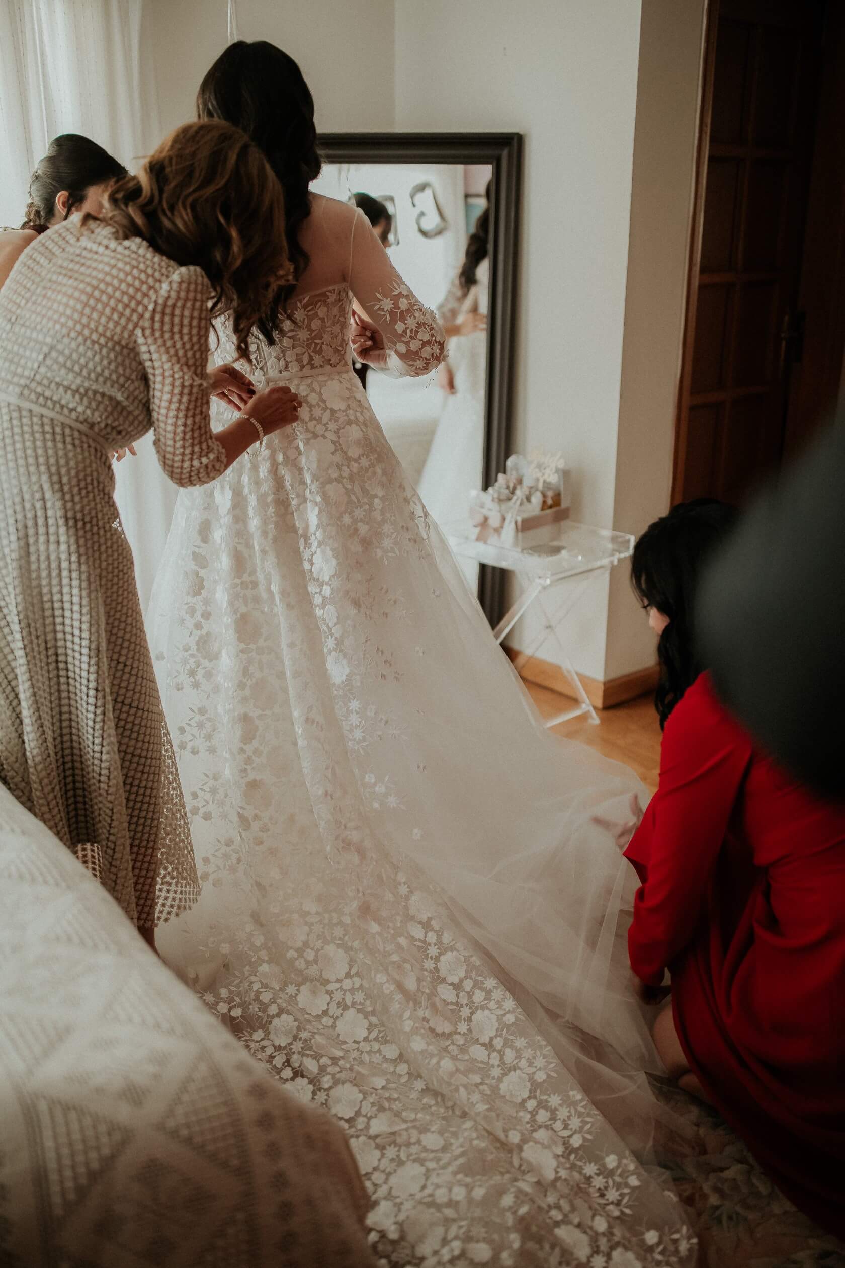 Bride’s dress adjusted by three women, mirror visible.