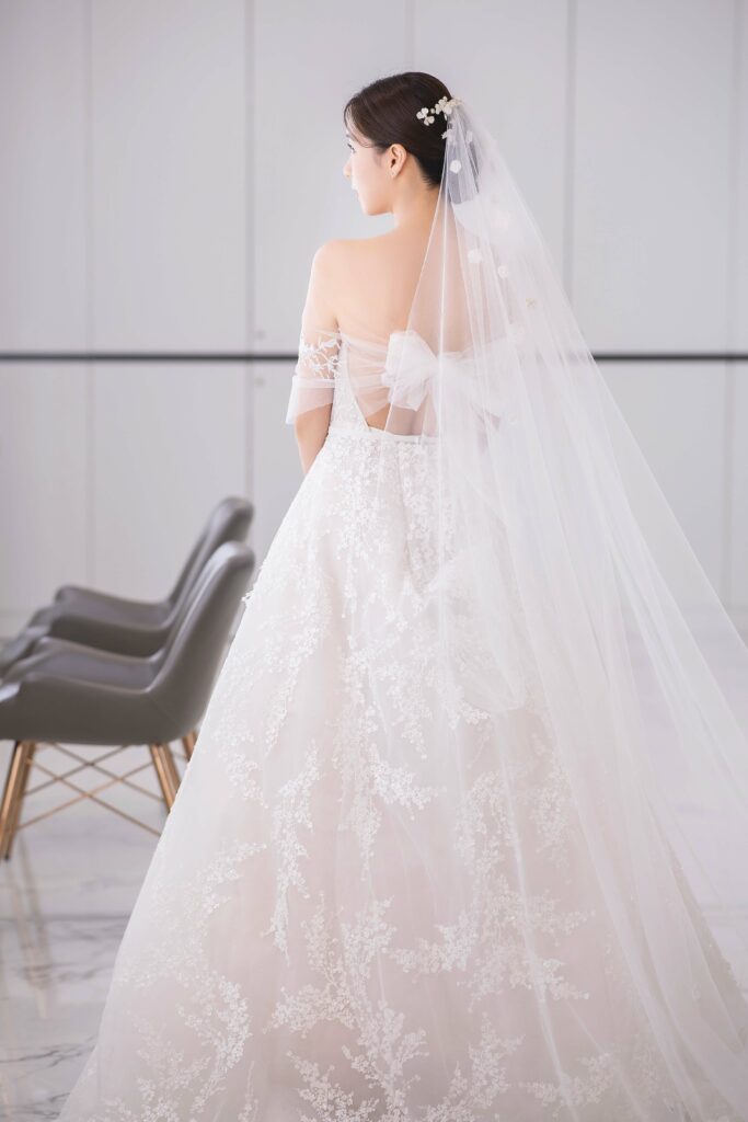 Bride in lace gown stands by black chairs.