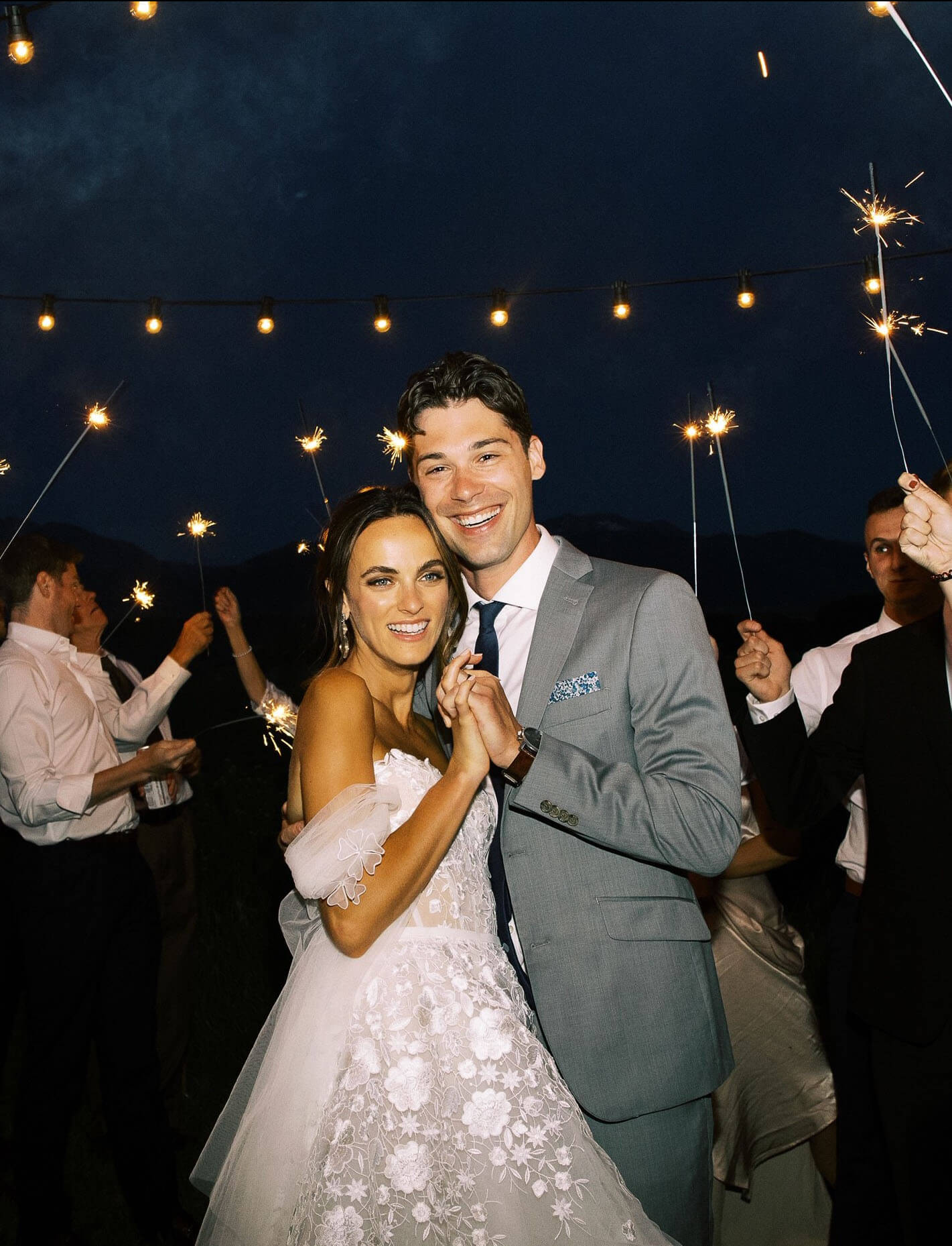 Bride and groom embrace under string lights, sparklers.
