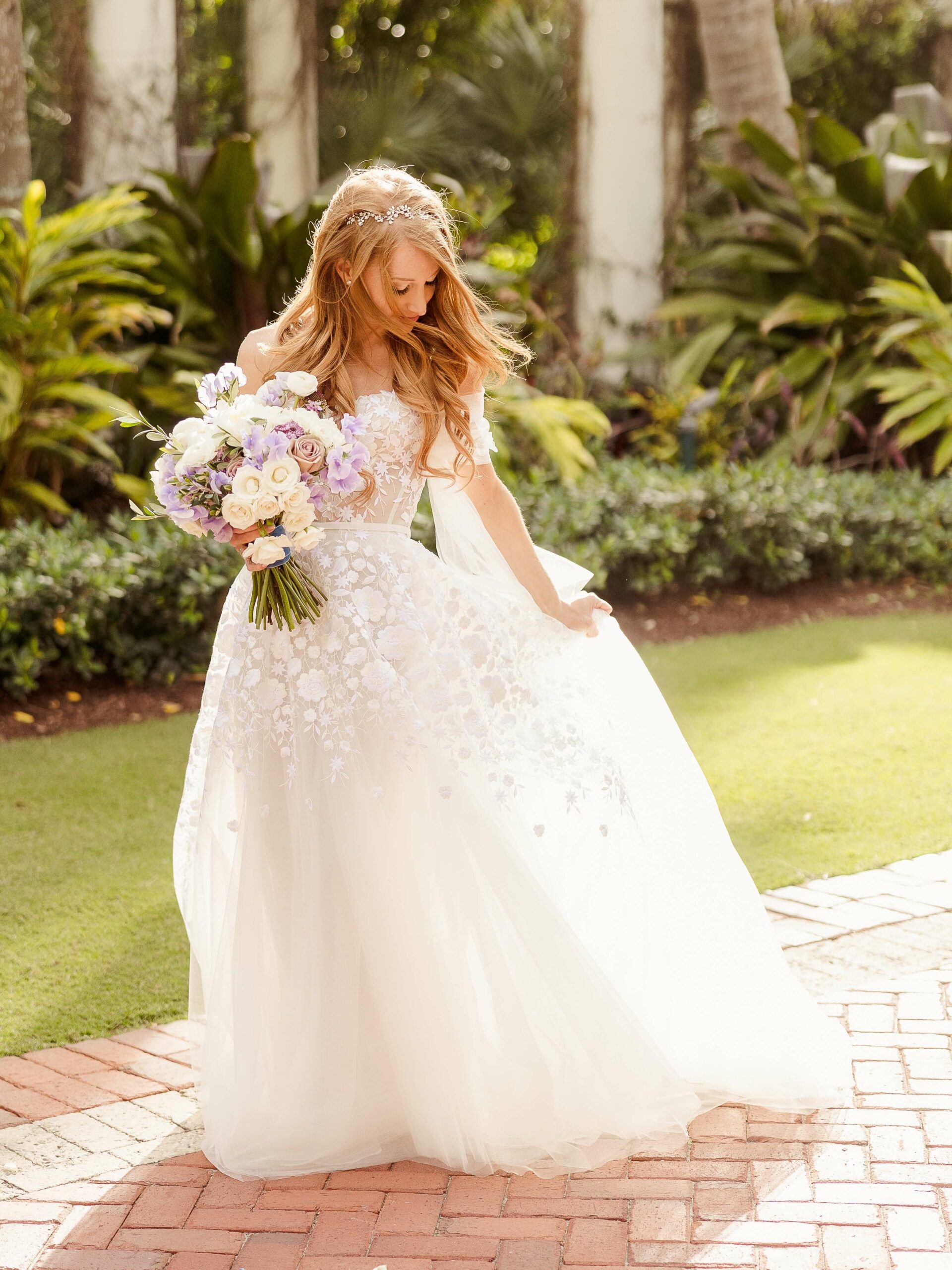 Bride smiling, holding bouquet on sunlit path.