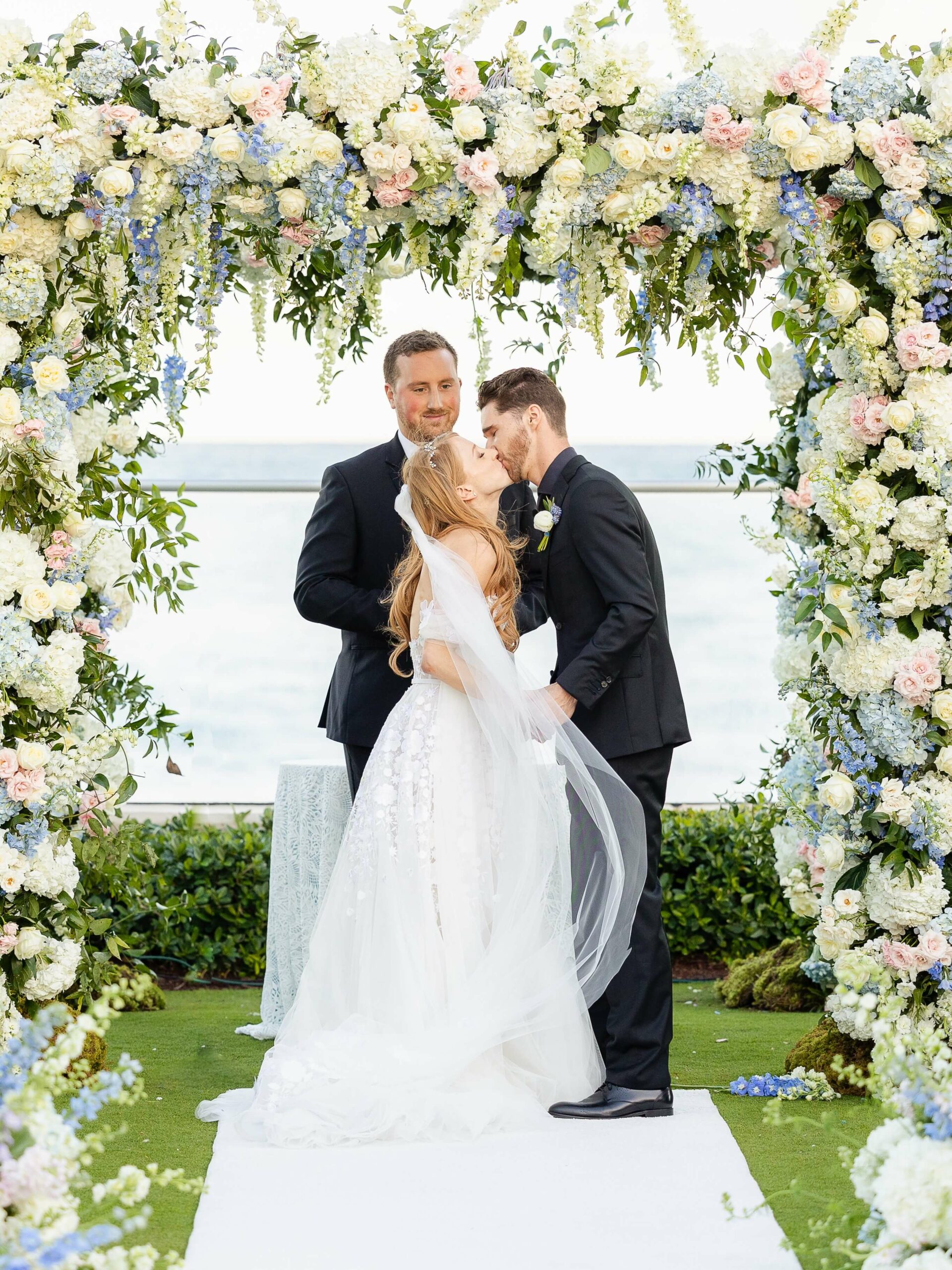 Bride and groom kiss under floral wedding arch.