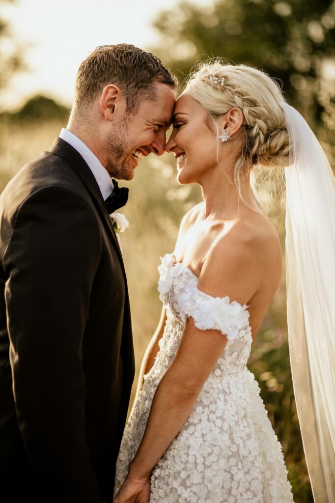 Bride and groom smiling, foreheads touching outdoors.
