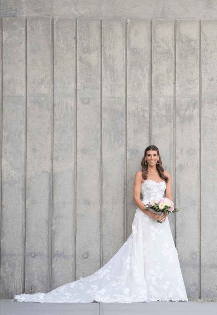 Smiling bride holding bouquet by concrete wall.