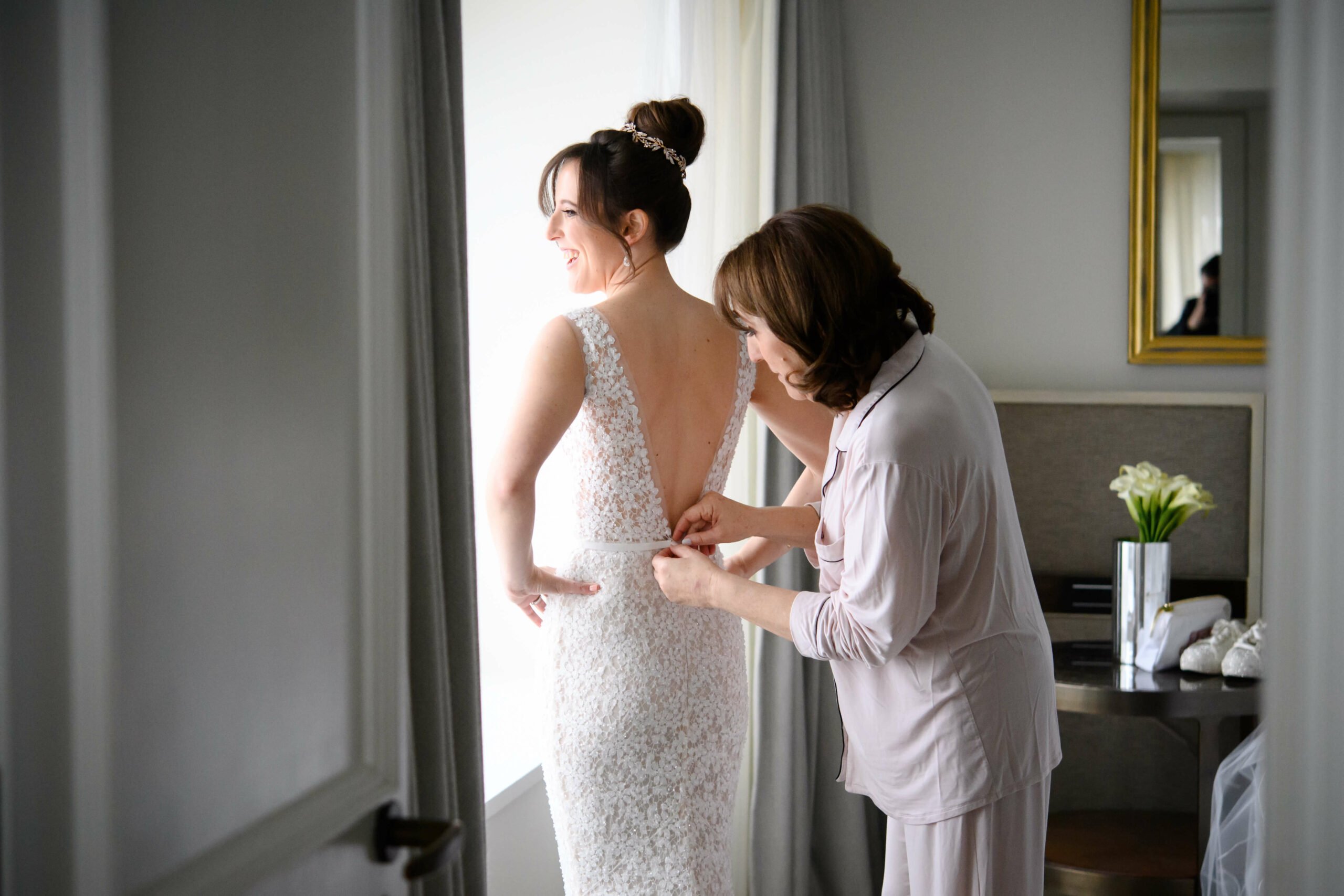 Woman helps bride adjust gown by window.