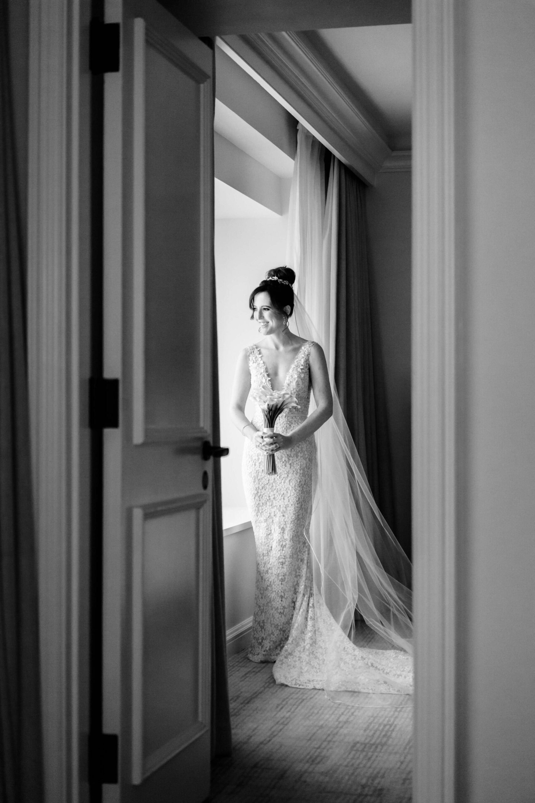 Bride in lace gown stands by sunlit window.