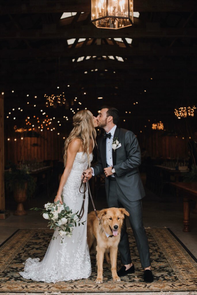 Bride and groom kiss with dog under lights.
