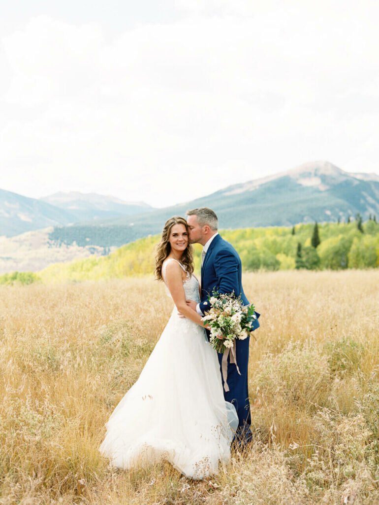 Groom kisses smiling bride in mountain field.