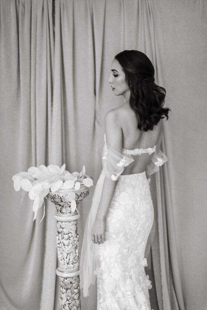 Bride in lace gown beside pedestal, black-and-white.