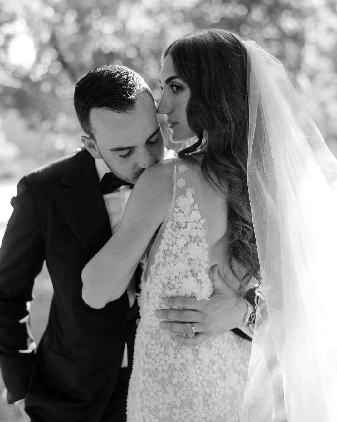 Groom kisses bride’s shoulder outdoors, soft light.