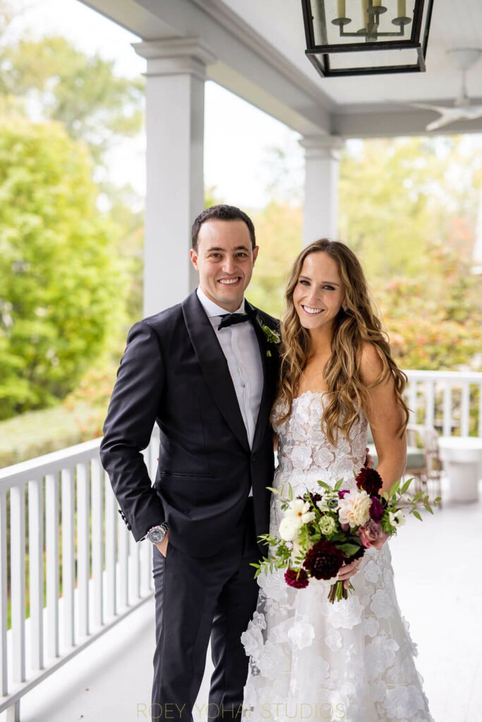 Smiling wedding couple on porch with bouquet.