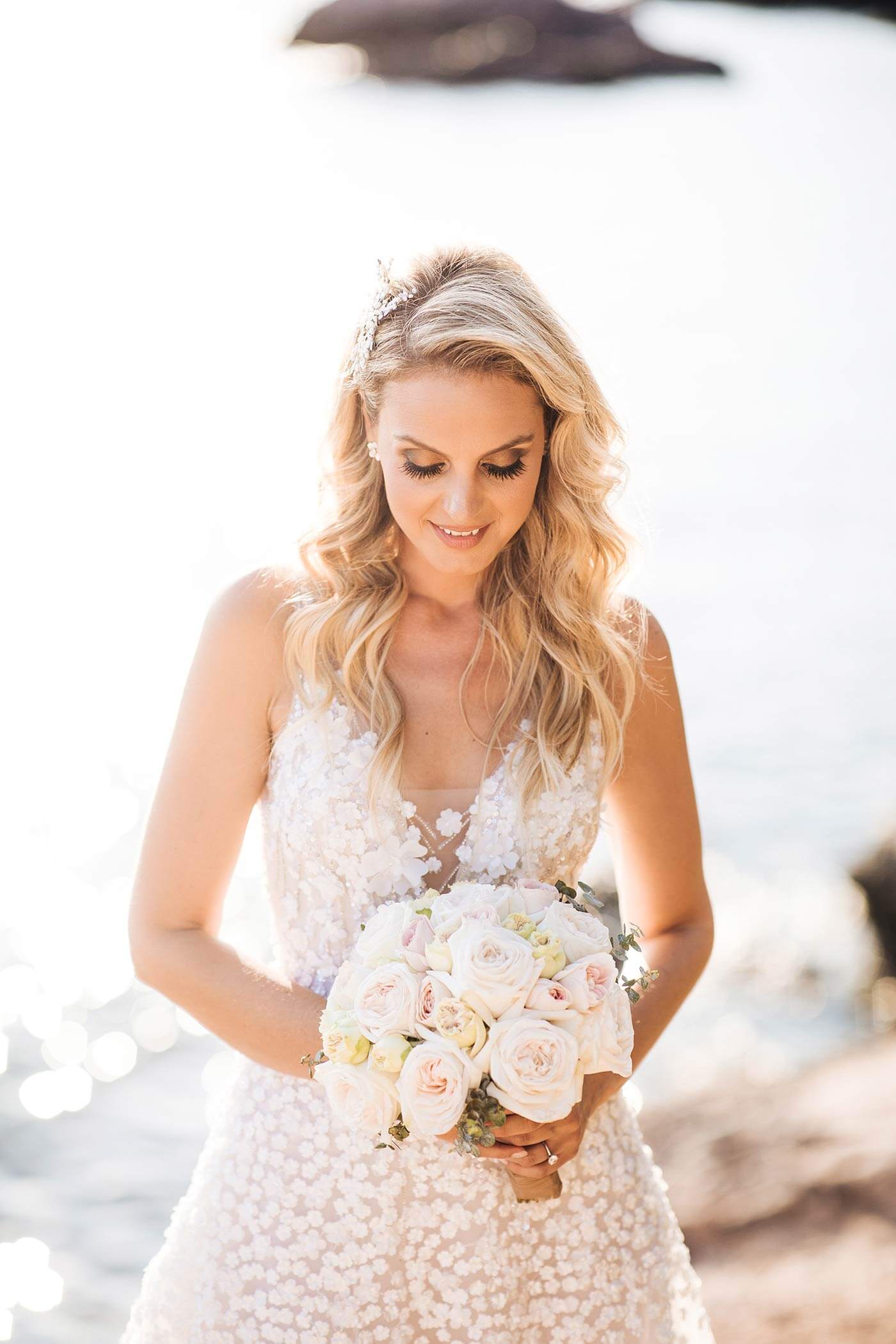 Bride in lace dress smiles by water, bouquet.
