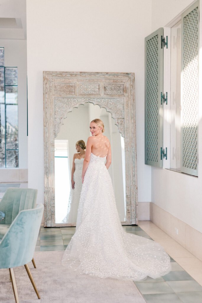 Bride in white gown before ornate mirror.