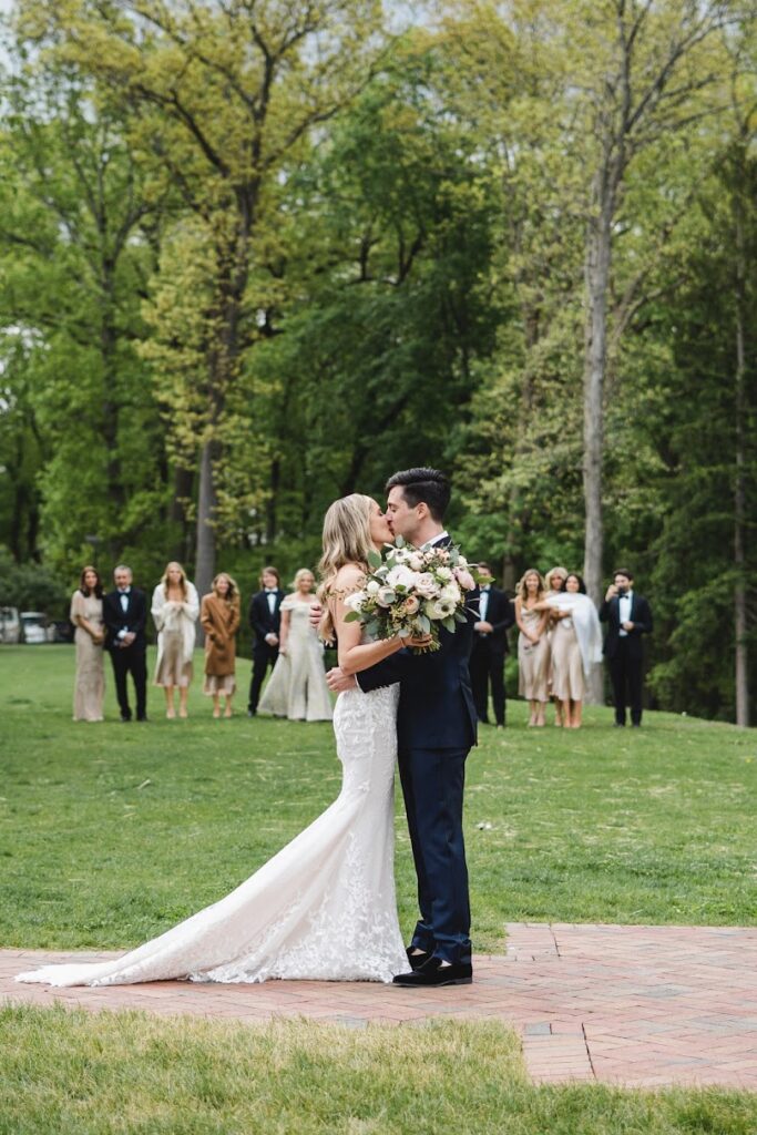 Bride and groom kiss on lawn, wedding party behind.