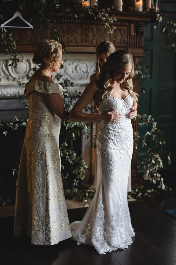 Brides dress adjusted by two women, fireplace.