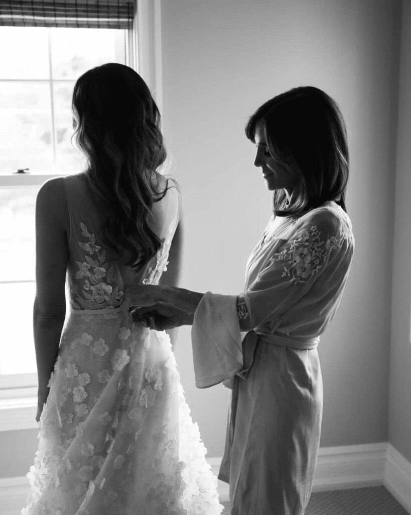 Woman helps bride button wedding dress, black-and-white.