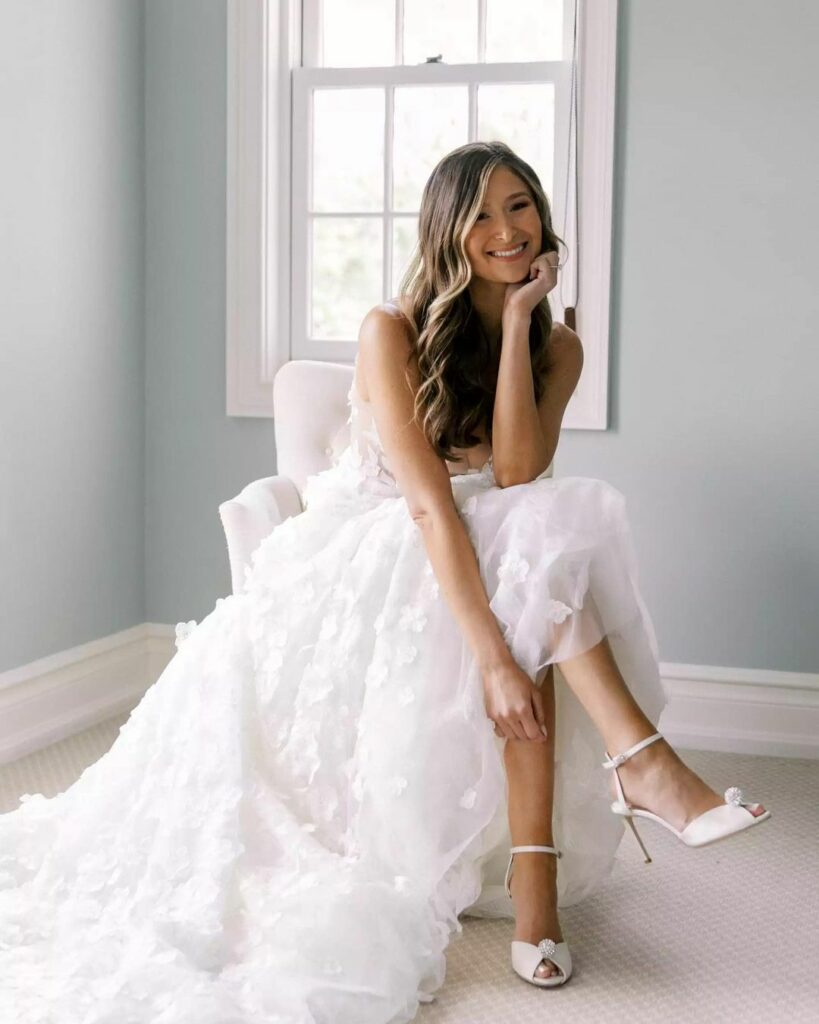 Smiling bride in white floral dress, seated.