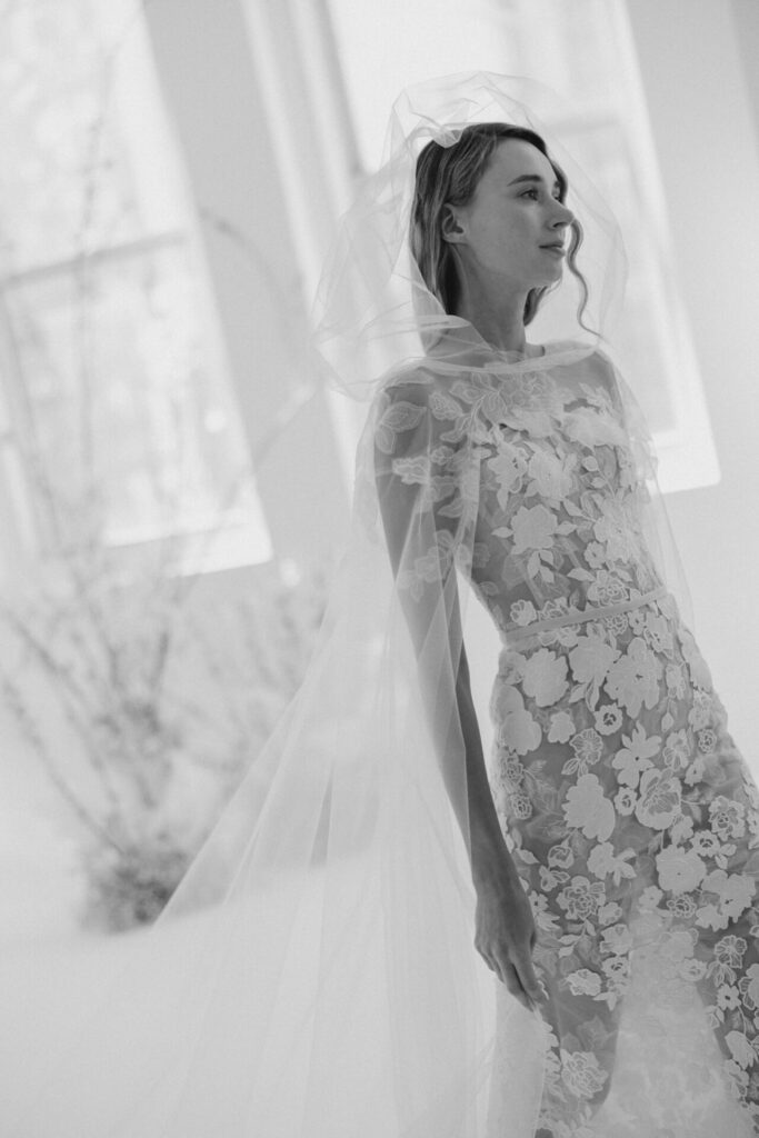 Smiling bride in lace gown by window.