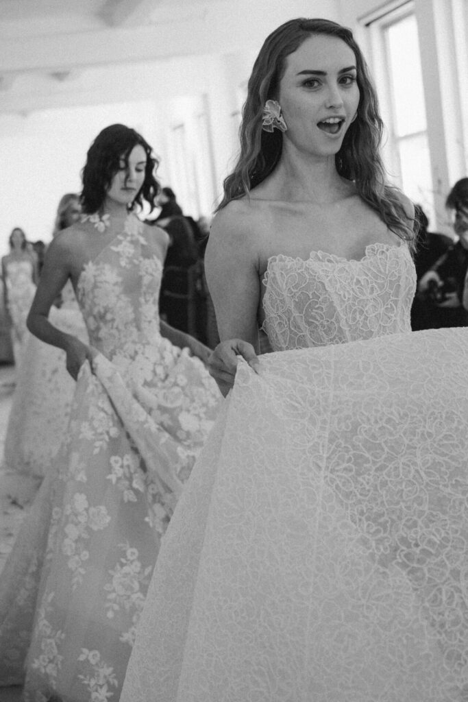 Two brides in lace gowns, people behind.