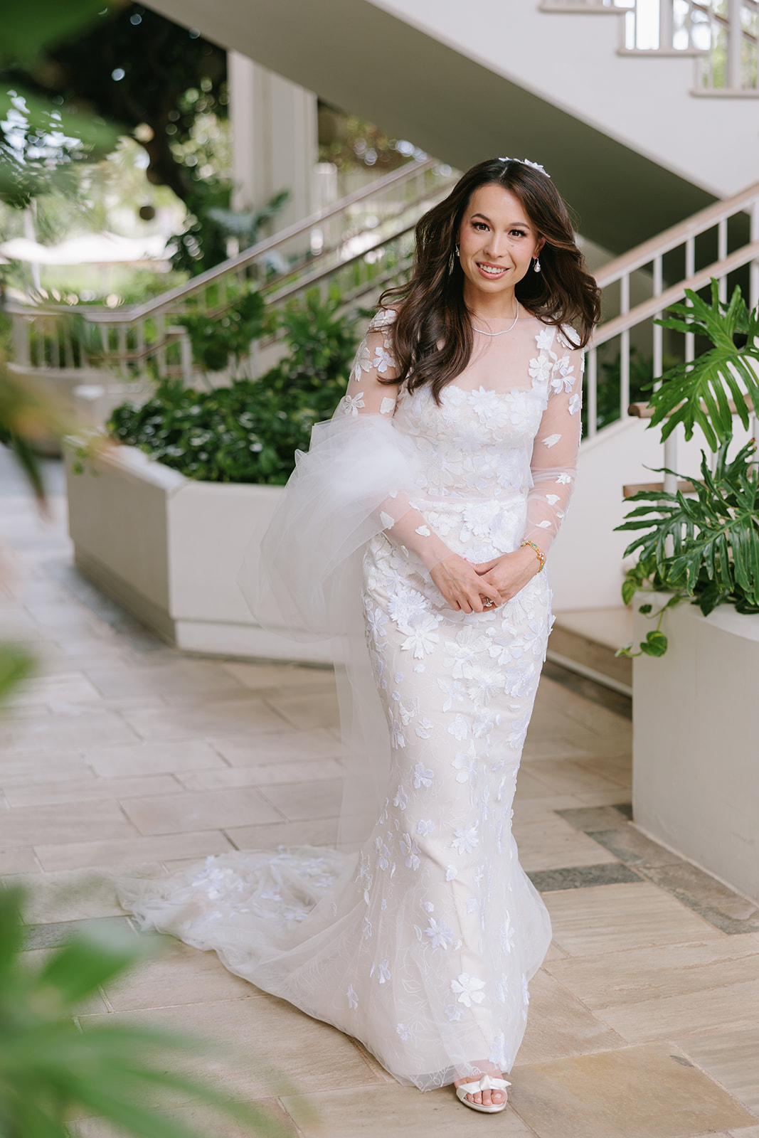 Bride in lace gown by plants and staircase.
