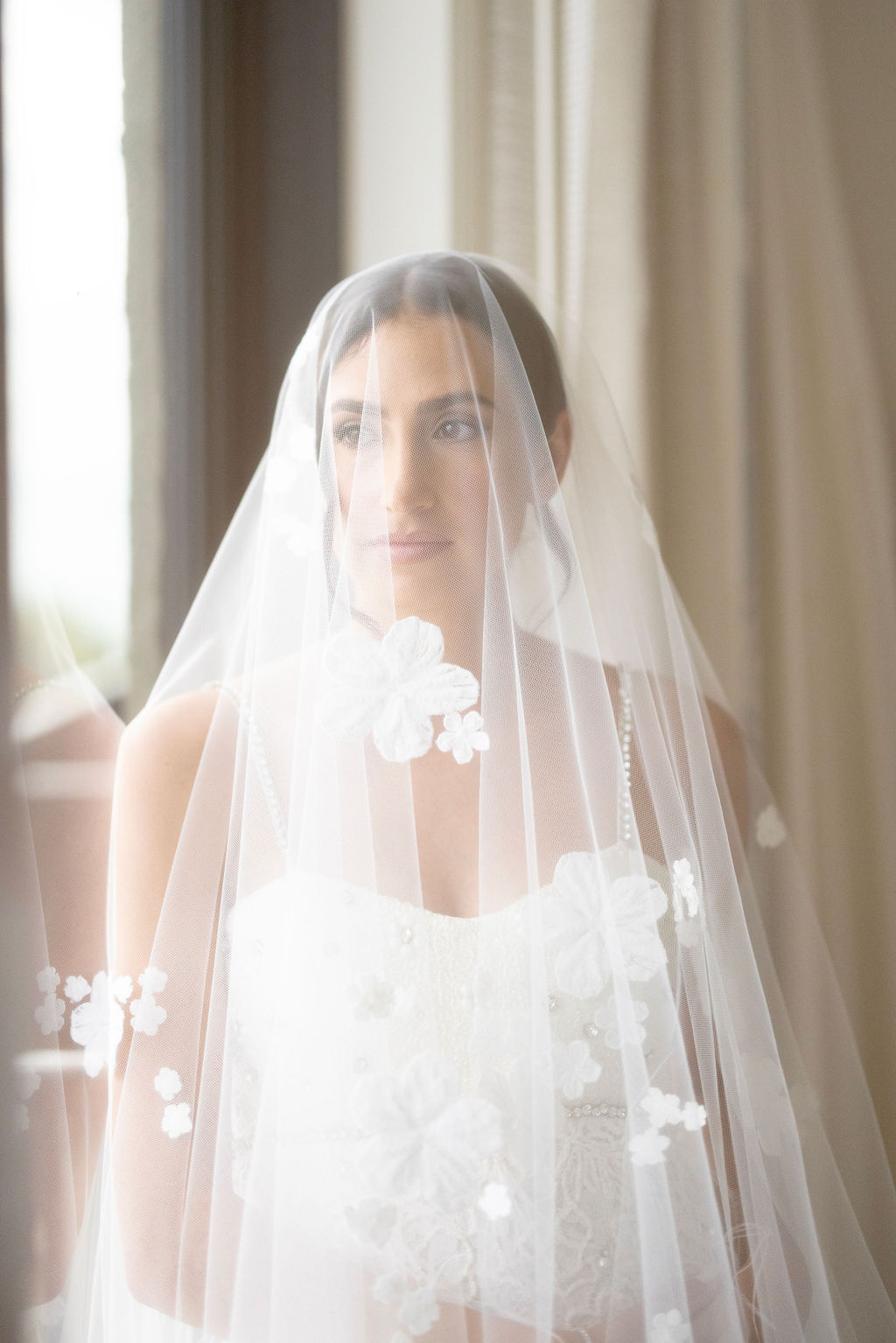 Bride in floral veil gazes out window.