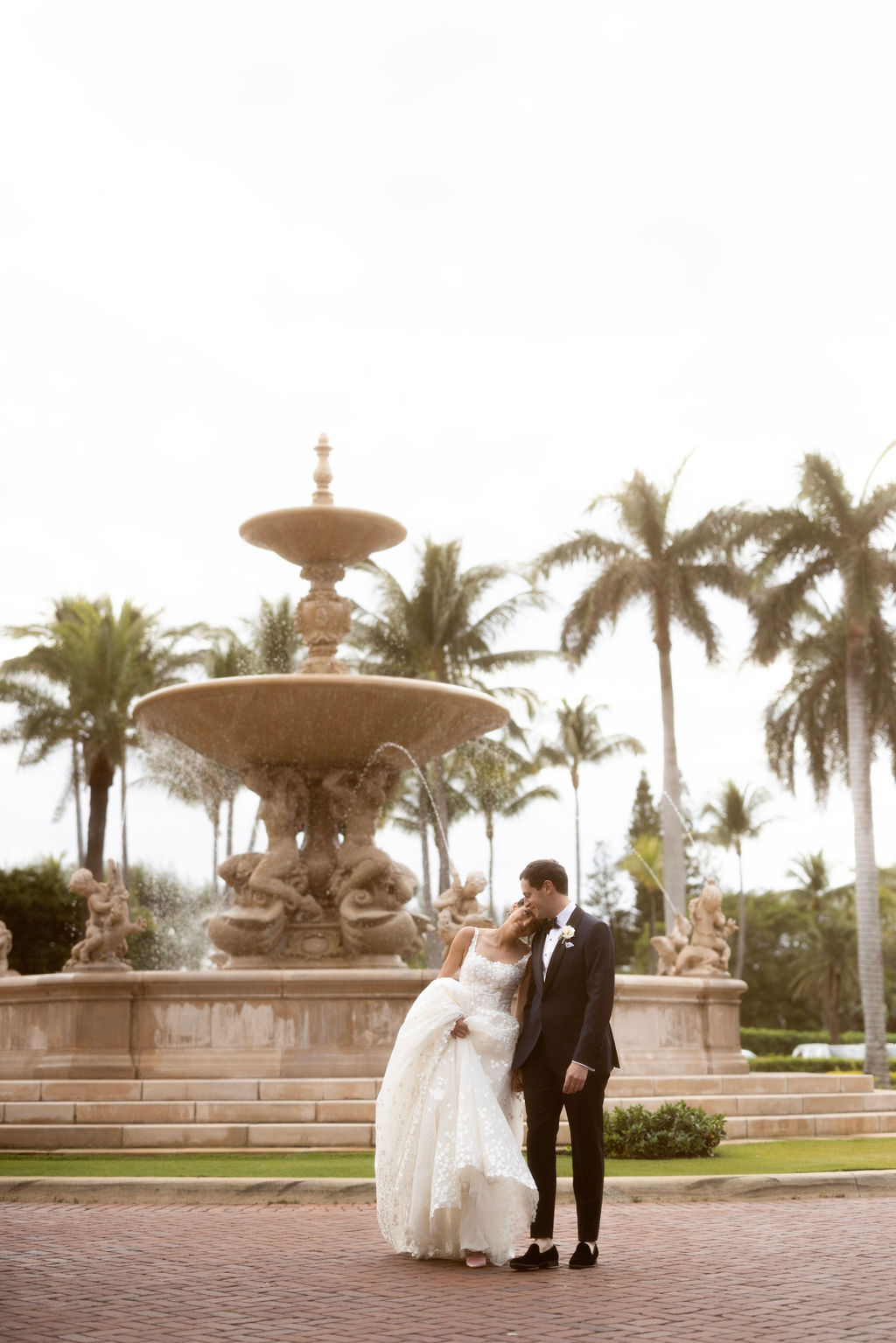 Bride and groom kiss by sunlit fountain.