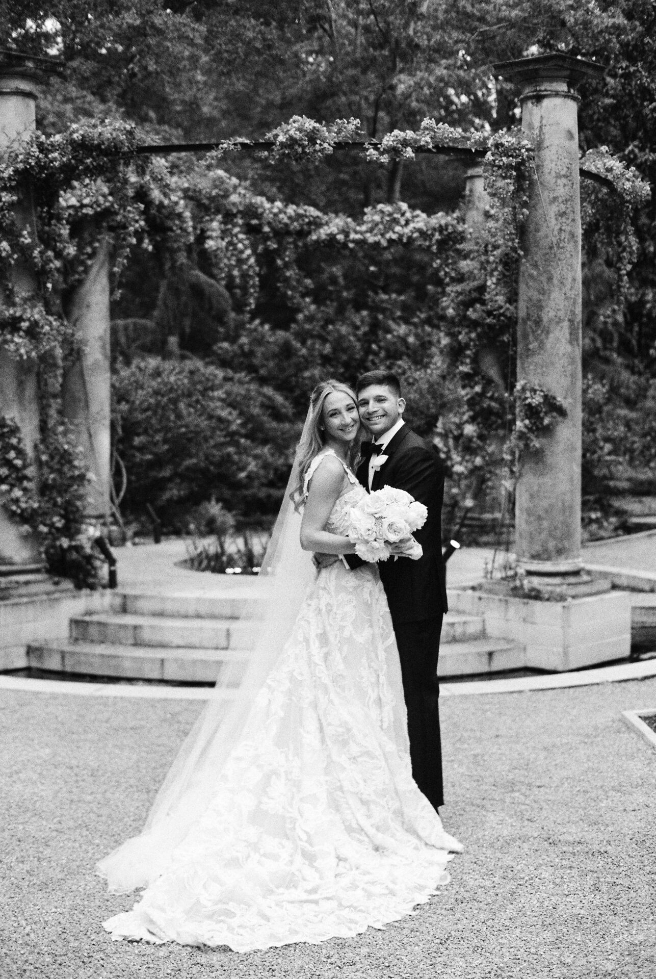 Bride and groom smiling outdoors amid greenery.