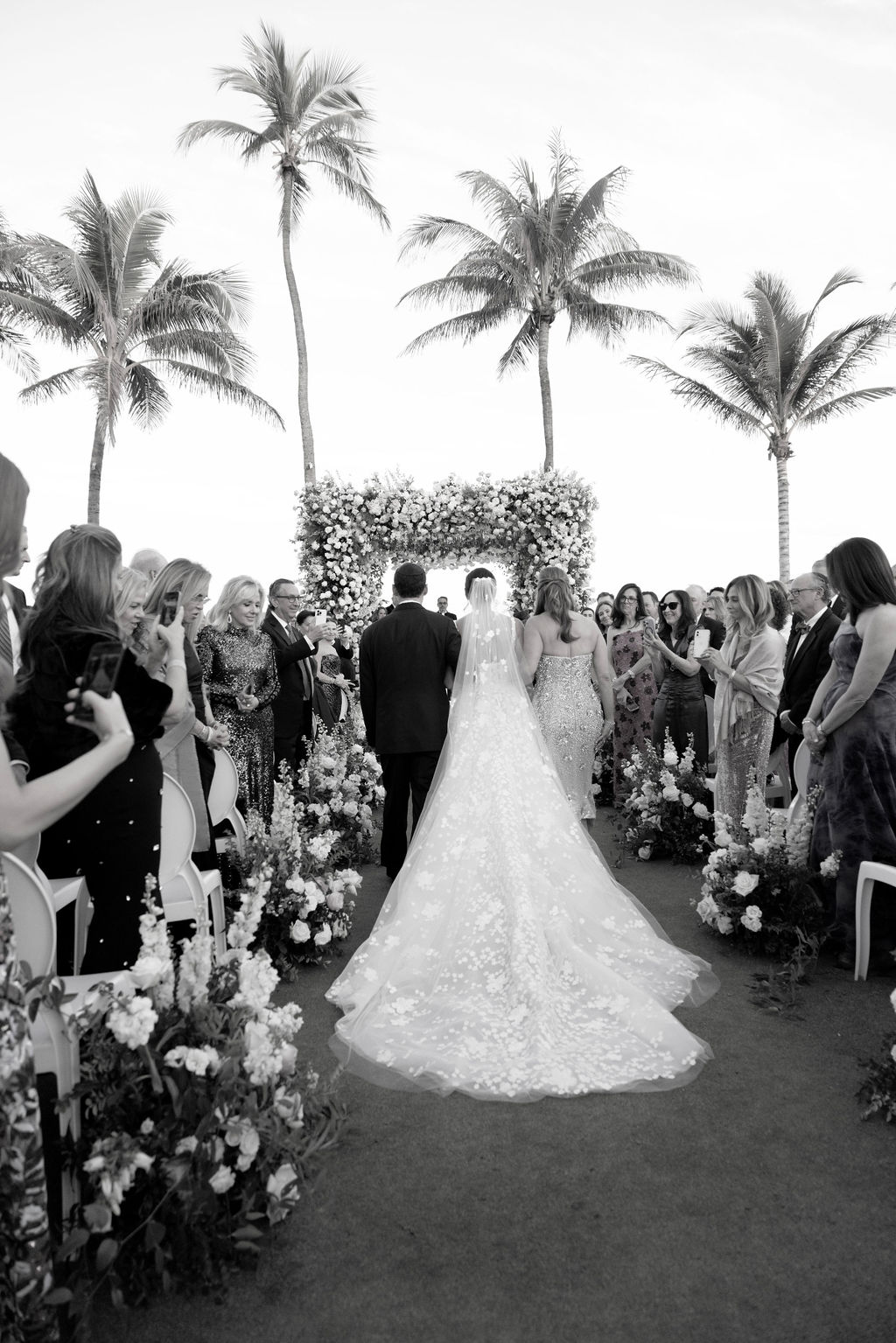 Bride walks aisle outdoors, escorted, black-and-white.