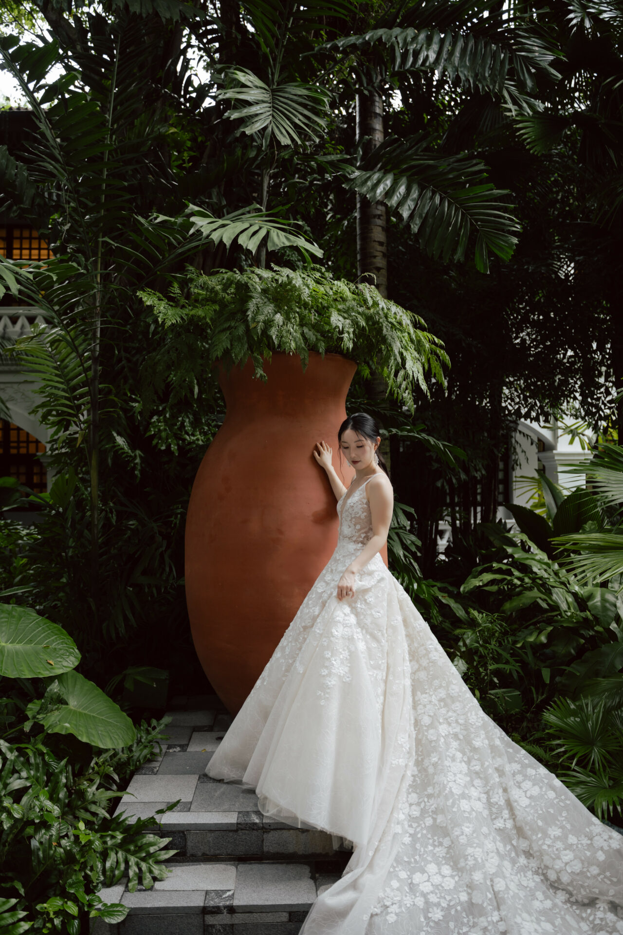 Bride in floral gown on steps, tropical plants.