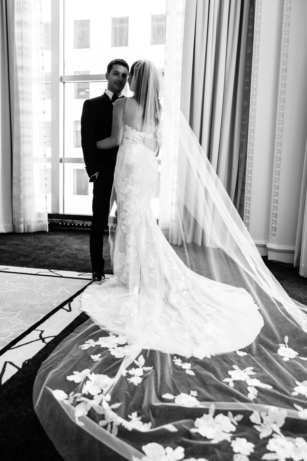 Bride and groom embrace by window, black-and-white.