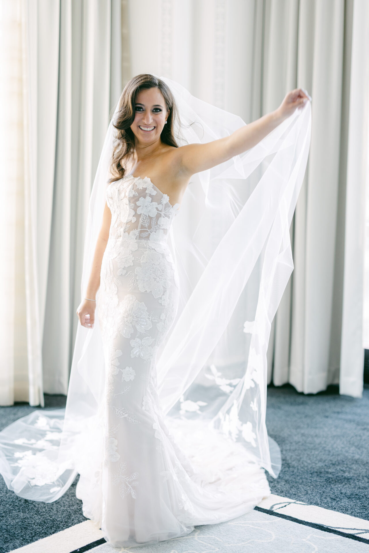 Bride in lace dress holds up veil indoors.