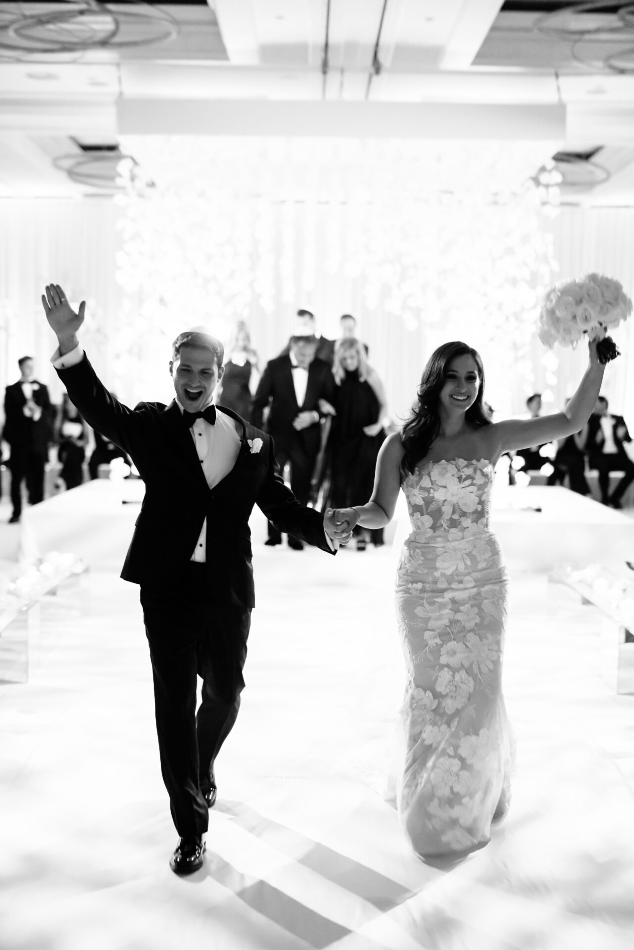 Smiling newlyweds walking aisle, guests seated, grayscale.