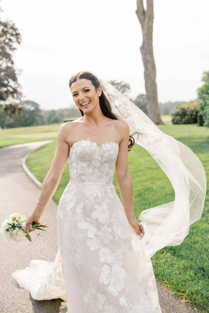 Smiling bride in lace dress holds bouquet outdoors.