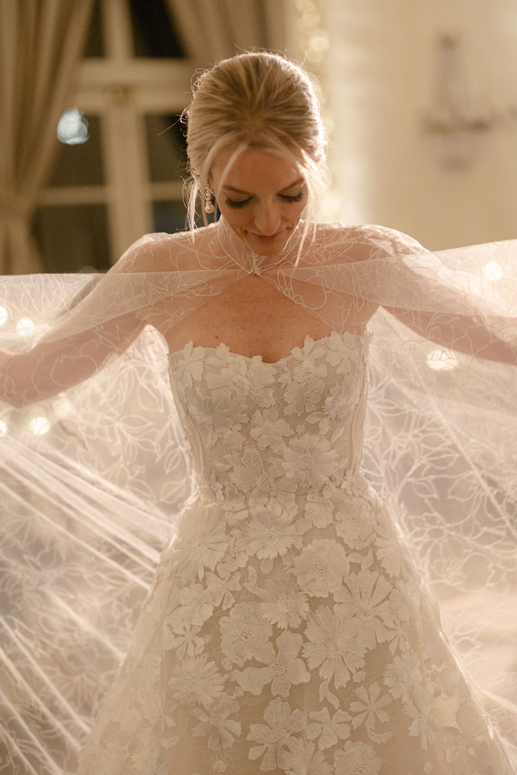 Bride in lace gown holds sheer embroidered veil.