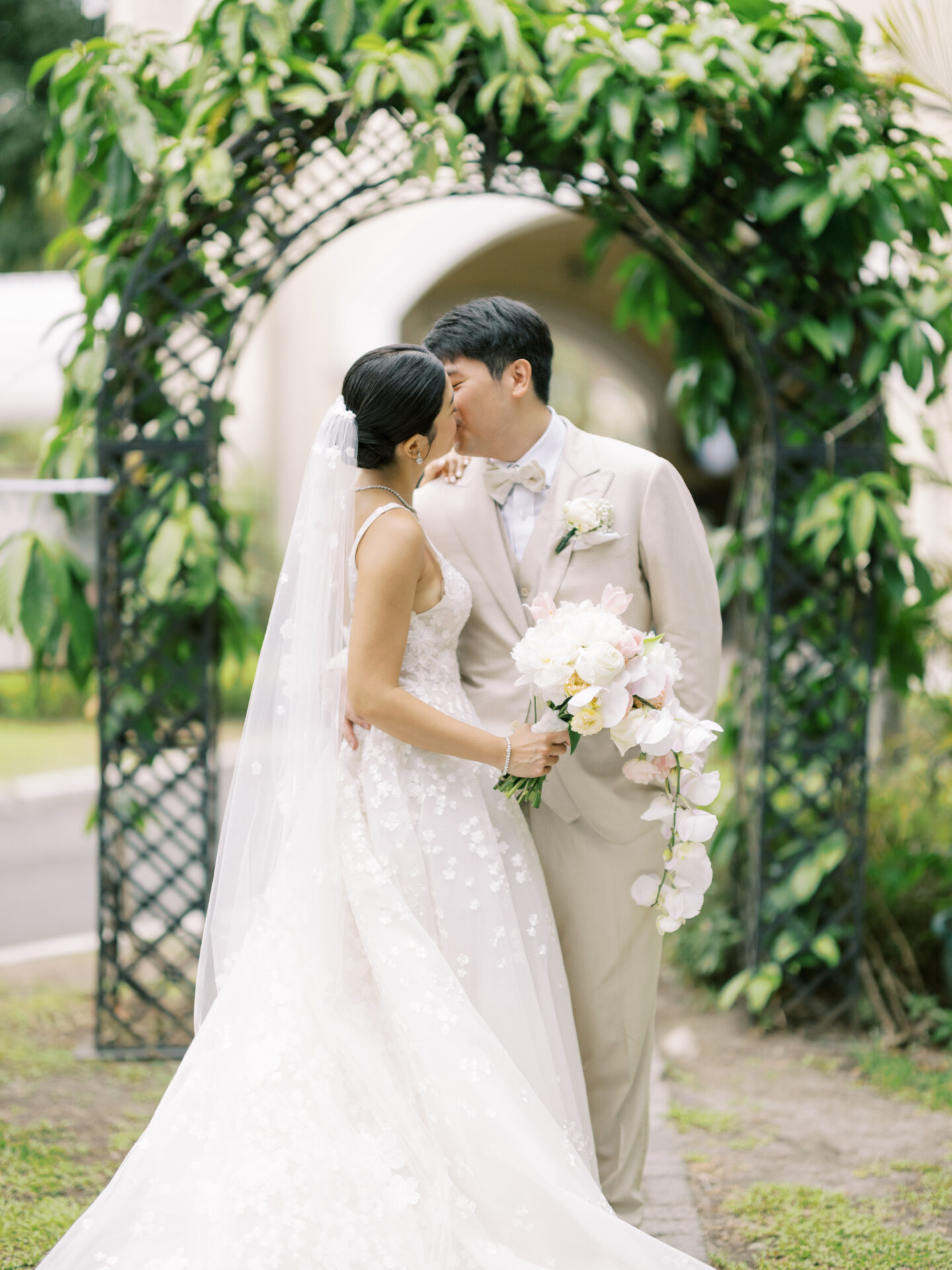Bride and groom kiss under green archway.