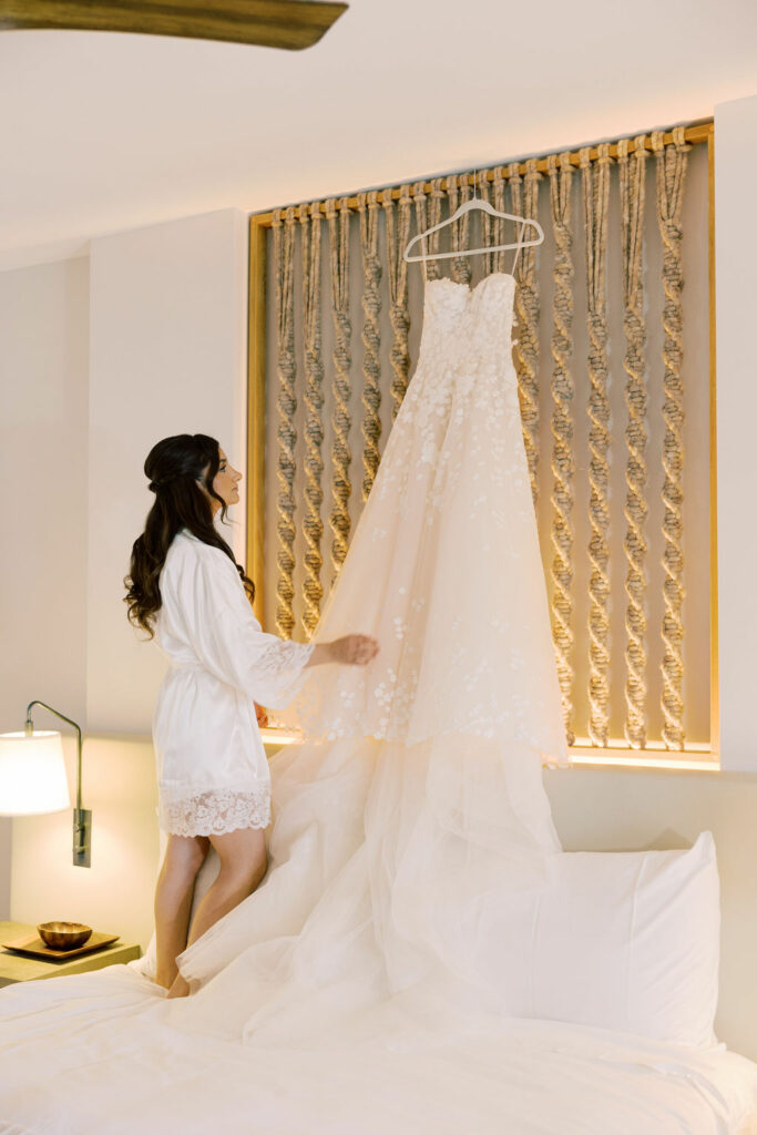 Bride in robe admires wedding gown above bed.