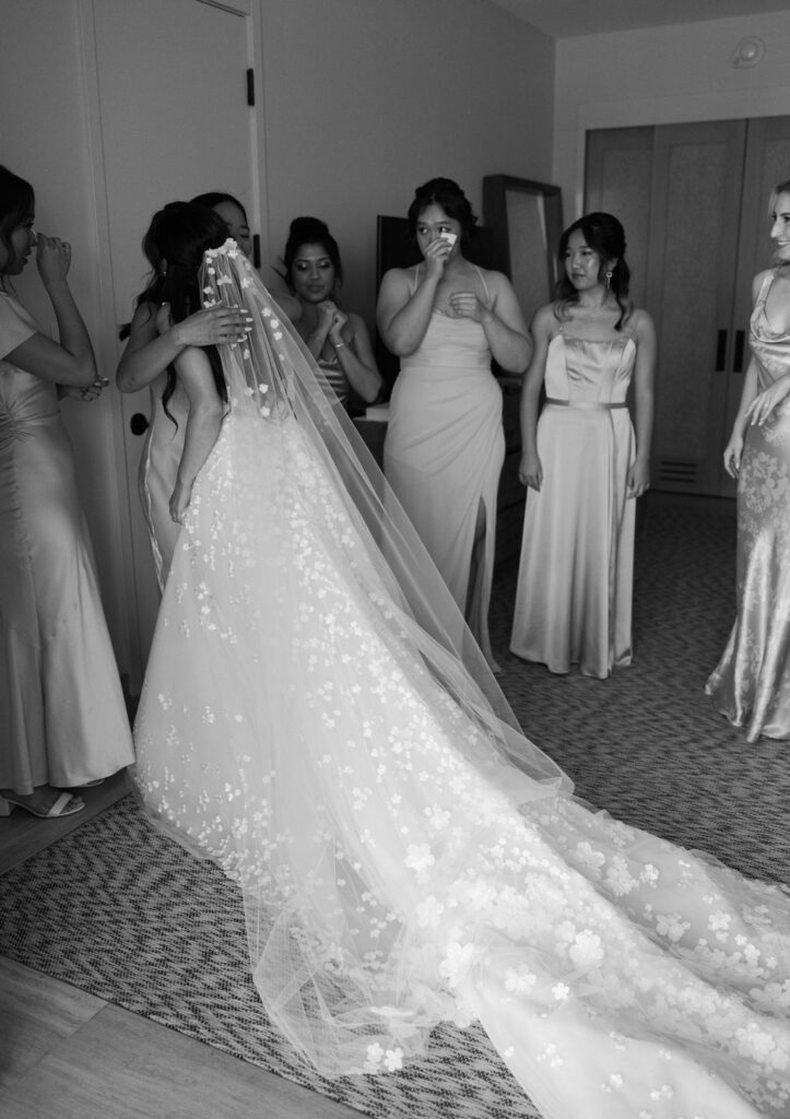 Bride hugs bridesmaid; others watch, emotional moment.