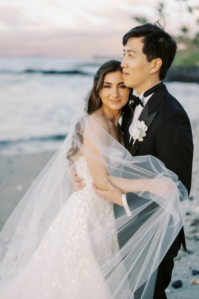Bride and groom embrace on beach at sunset.