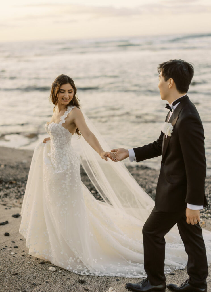 Bride and groom in couture walk on beach.