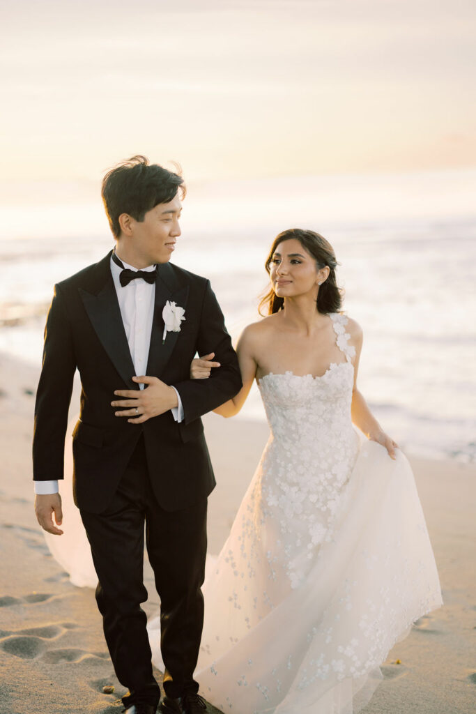 Bride and groom walk on beach at sunset.