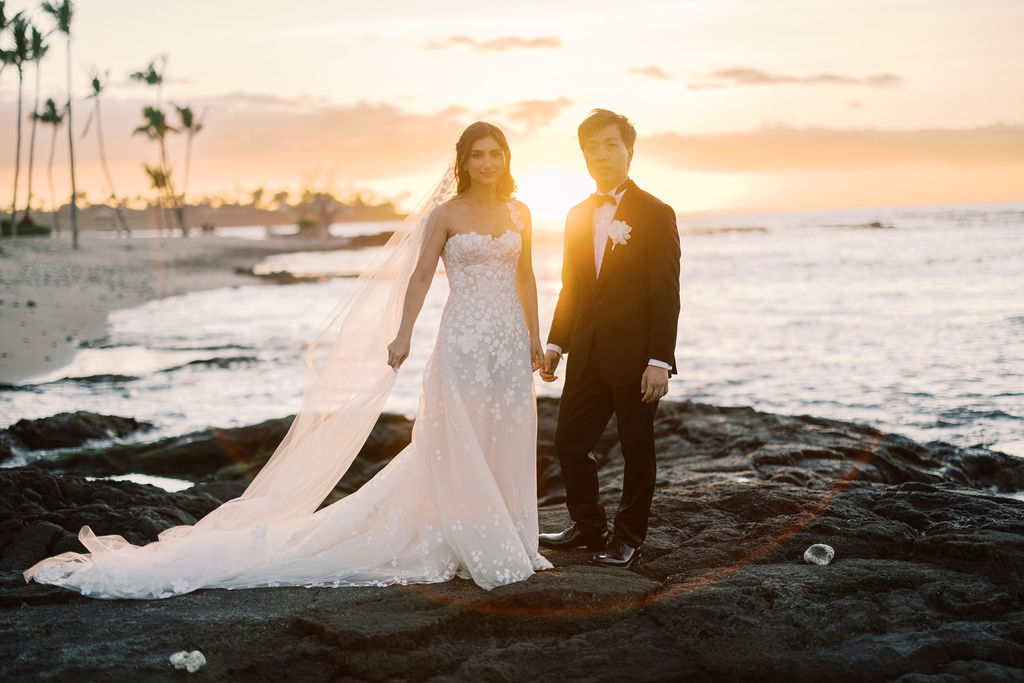 Bride and groom hold hands on rocky shore.