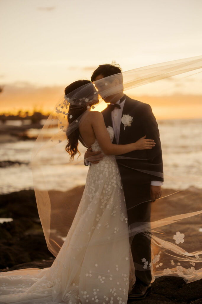 Bride and groom kiss under veil at sunset.