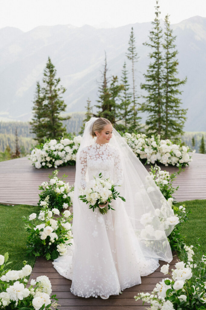 Bride in lace gown outdoors with white flowers.