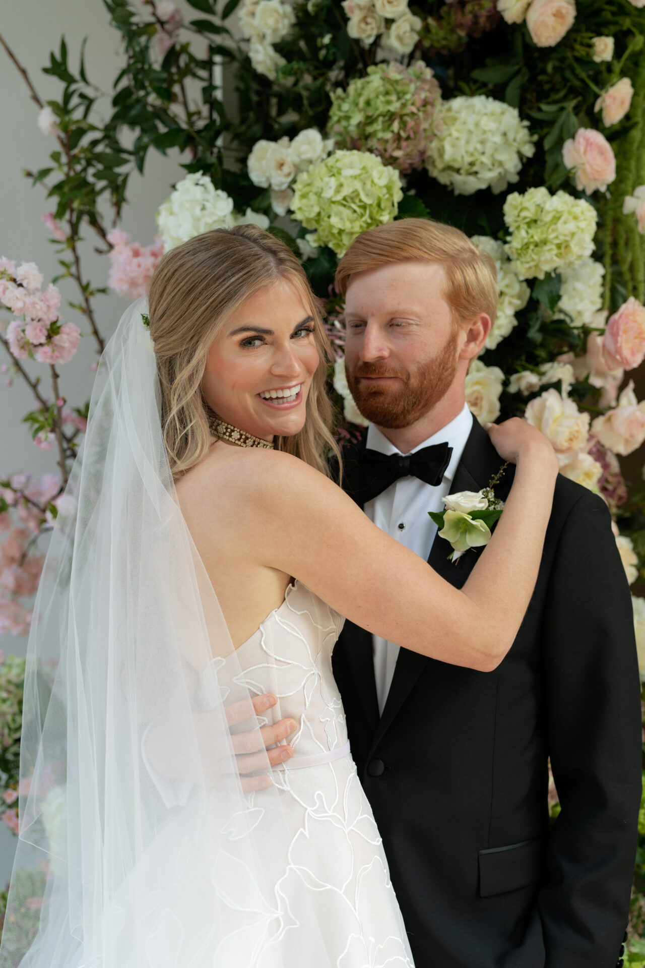 Bride and groom embrace before floral arrangement.