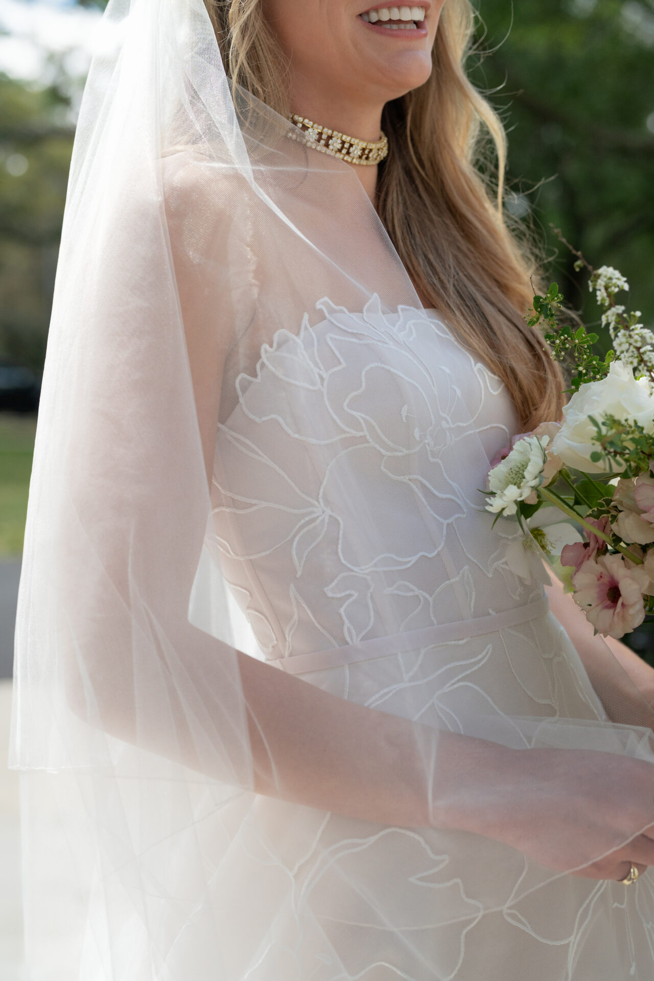 Smiling bride holds bouquet outdoors.