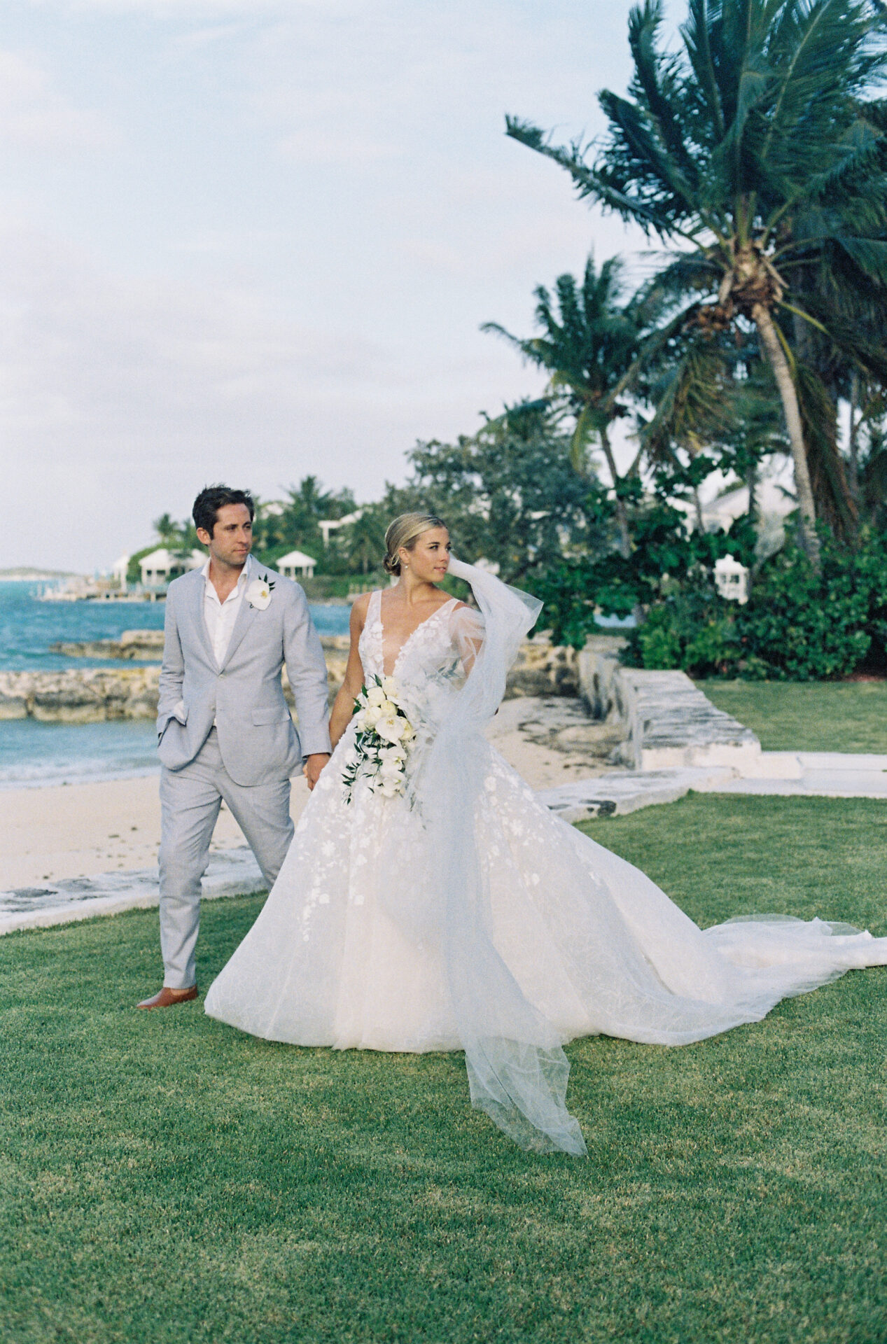 Bride and groom walk on lawn by ocean.