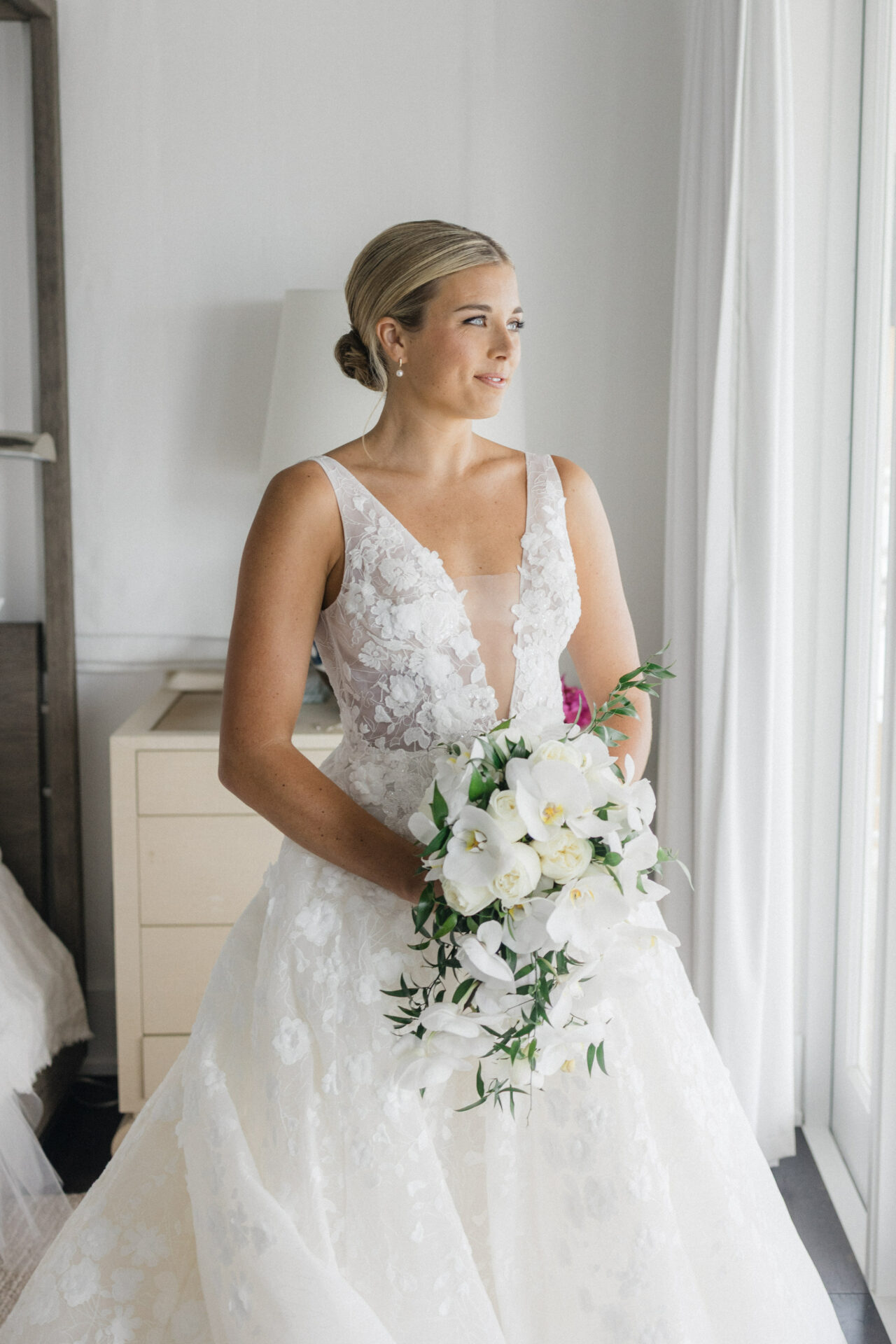 Bride in lace dress gazes out window.