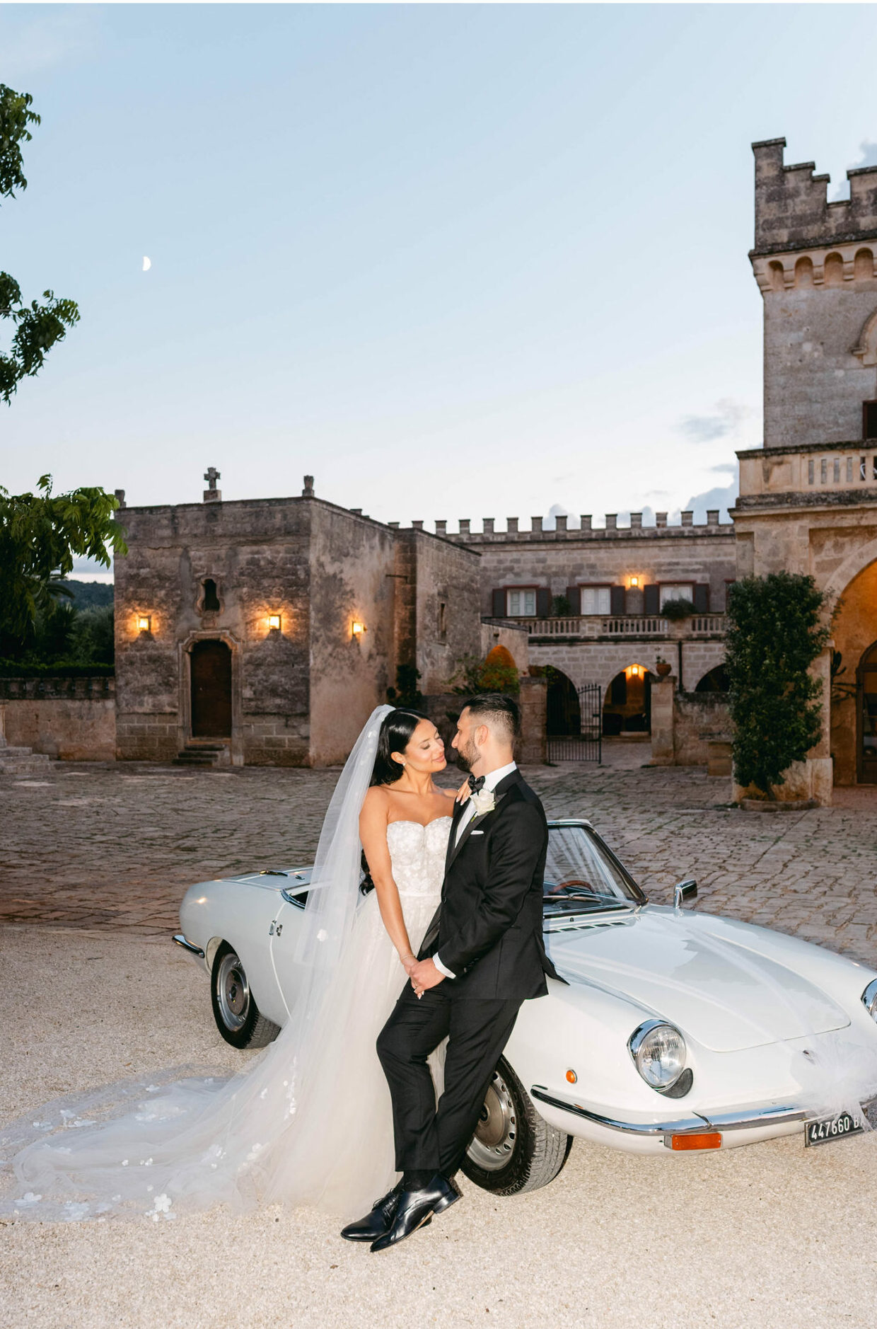 Bride and groom embrace by vintage car at sunset.